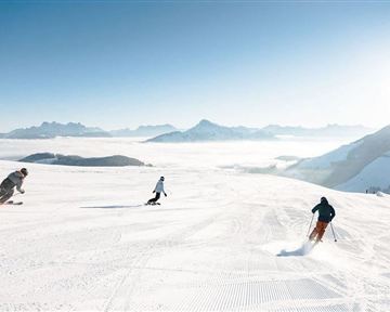A sunny day on the ski slope with three skiers. The untouched, snow-covered landscape extends to the horizon.