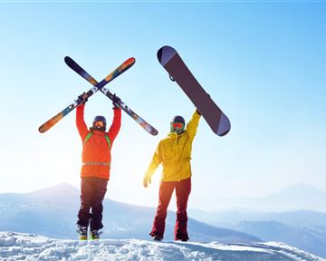 Zwei Personen stehen auf einem schneebedeckten Berggipfel. Eine Person hält Skier hoch, während die andere ein Snowboard hebt.