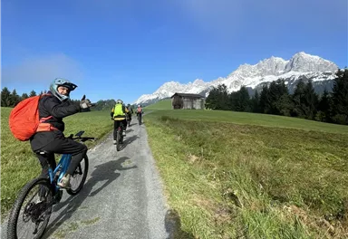 Eine Gruppe von Radfahrern fährt auf einem Weg durch die grüne Landschaft. Im Hintergrund sind schneebedeckte Berge und ein blauer Himmel zu sehen.