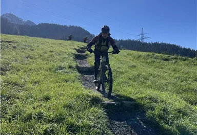 Ein Radfahrer fährt einen schmalen Weg auf einer grünen Wiese. Im Hintergrund sind Berge und ein klarer blauer Himmel sichtbar.
