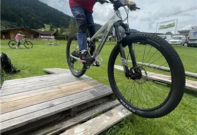 Ein Junge fährt mit dem Fahrrad über eine Holzrampe. Im Hintergrund sind weitere Radfahrer und eine grüne Wiese zu sehen.
