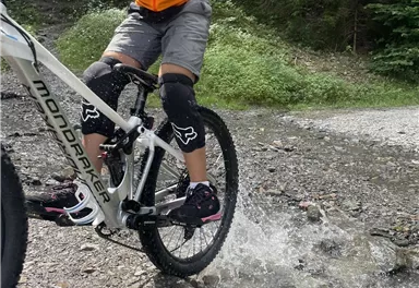 Ein Fahrradfahrer springt durch eine Pfütze auf einem Waldweg. Er trägt einen orangefarbenen Trikot und Schutzkleidung.
