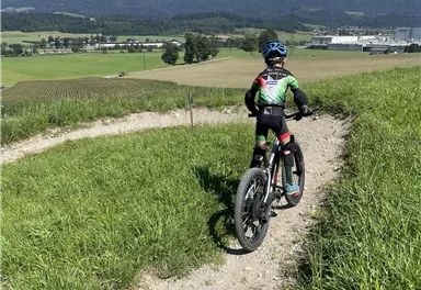 Ein Junge fährt mit einem Fahrrad einen Weg durch eine grüne Landschaft. Im Hintergrund sind Berge und ein blauer Himmel zu sehen.