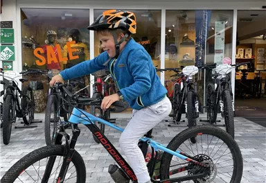 Ein Junge fährt auf einem Mountainbike vor einem Sportgeschäft. Er trägt einen Helm und hat viel Spaß beim Radfahren.