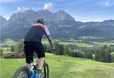 Ein Radfahrer fährt auf einem grünen Hügel mit Blick auf majestätische Berge. Der Himmel ist klar und blau, ideale Bedingungen für Outdoor-Aktivitäten.