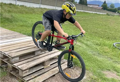 Ein Junge fährt mit einem Fahrrad über eine Holzkonstruktion. Er trägt einen Helm und konzentriert sich auf die Fahrt im Freien.