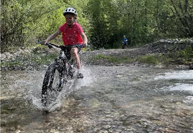 Ein Kind fährt mit dem Fahrrad durch ein Gewässer und spritzt dabei Wasser. Im Hintergrund sind Bäume und ein weiterer Radfahrer zu sehen.