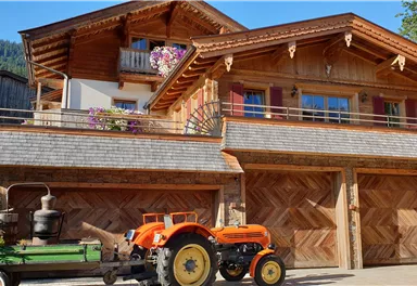 Ein traditionelles Holzhaus mit Balkonen und bunten Blumen. Davor steht ein orangefarbener Traktor auf einem geschmackvollen Hof.