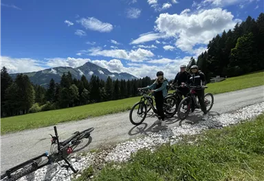 Drei Radfahrer stehen auf einem Weg in einer grünen Landschaft mit Bergen im Hintergrund. Der Himmel ist blau mit einigen Wolken.