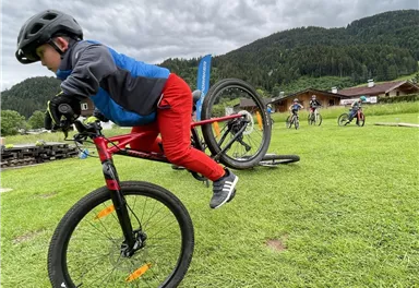 Ein Junge fährt mit seinem Fahrrad über eine kleine Rampe. Im Hintergrund sind weitere Kinder und eine grüne Landschaft zu sehen.