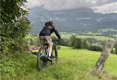 Ein Mountainbiker fährt durch eine grüne Wiese mit Bergen im Hintergrund. Der Himmel ist bewölkt und die Landschaft wirkt ruhig und einladend.