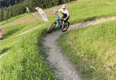 Ein Radfahrer fährt auf einem schmalen Pfad inmitten von grünen Wiesen und Bäumen. Im Hintergrund sind einige Bergbahn-Stützen zu sehen.