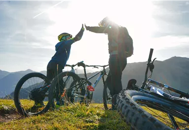 Ein Vater und sein Sohn geben sich auf einem Berg einen High-Five. Im Hintergrund sind Mountainbikes und eine wunderschöne Berglandschaft zu sehen.
