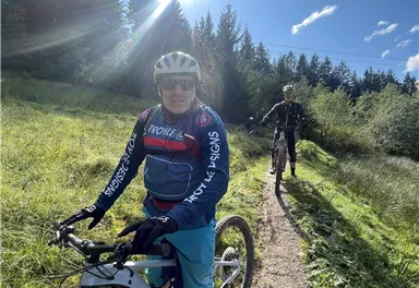 Zwei Männer fahren mit Mountainbikes auf einem schmalen Weg durch eine grüne Landschaft. Die Sonne scheint und der Himmel ist teilweise bewölkt.
