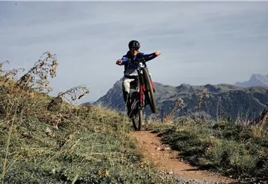 Ein junger Radfahrer fährt auf einem schmalen Pfad durch eine bergige Landschaft. Im Hintergrund sind sanfte Hügel und ein klarer Himmel zu sehen.