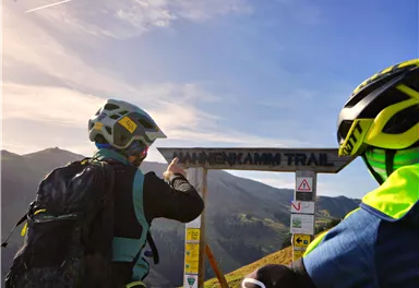 Zwei Mountainbiker stehen vor einem Schild, das den "Bahnenkammt Trail" anzeigt. Die Umgebung ist bergig mit klarem Himmel und Sonnenlicht.