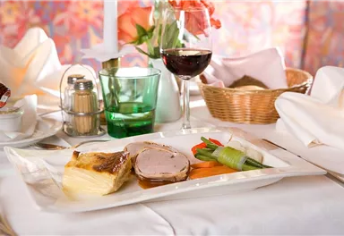 Ein elegantes Gericht mit Fleisch, Gemüse und Kartoffeln auf einem weißen Teller. Dazu ein Glas Rotwein und dekorierte Tischelemente.