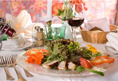 Ein frisch zubereitetes Gericht mit Hähnchenfilet, buntem Salat und Tomatenscheiben. Im Hintergrund sind ein Glas Rotwein und eine dekorative Tischdekoration zu sehen.