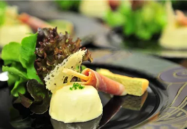 Ein stilvolles Gericht mit cremigem Hauptbestandteil, umgeben von frischem Salat und Schinken. Die Anrichtung auf einem eleganten schwarzen Teller sorgt für eine ansprechende Präsentation.