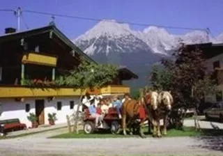 Ein charmantes Landhaus in den Bergen, umgeben von grüner Wiese und beeindruckenden Gipfeln. Im Vordergrund stehen Pferde mit einer Kutsche, die bereit zur Abfahrt sind.