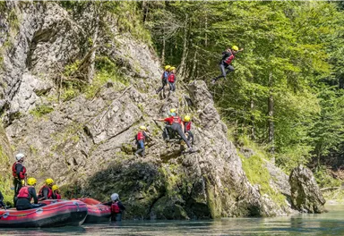 Eine Gruppe von Personen springt von einem Felsen ins Wasser. Im Hintergrund sind Bäume und ein Boot zu sehen.