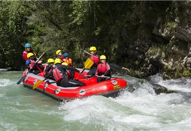 Eine Gruppe von Menschen fährt in einem roten Raft über einen wilden Fluss. Alle tragen Helme und Schwimmwesten und scheinen viel Spaß zu haben.