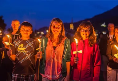 Eine Gruppe von Kindern und Erwachsenen hält Fackeln in der Dämmerung. Sie lächeln und scheinen an einer festlichen Veranstaltung teilzunehmen.