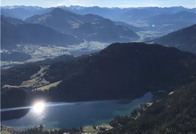 Eine atemberaubende Aussicht auf Berge und einen glitzernden See im Tal. Der Himmel ist klar und die Landschaft ist mit üppigem Grün bedeckt.