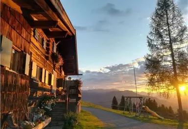 Ein malerisches Berghaus mit Holzwänden und bunten Pflanzen. Im Hintergrund ist ein Sonnenuntergang über den Bergen zu sehen.