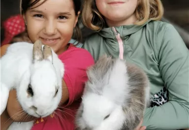 Zwei Mädchen halten liebevoll Kaninchen in ihren Armen. Sie lächeln glücklich in die Kamera.