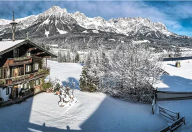 Eine malerische Winterlandschaft mit schneebedeckten Bergen und einem traditionellen Haus. Der Himmel ist klar und die Umgebung wirkt ruhig und einladend.