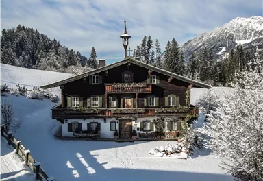 Ein pittoreskes Chalet umgeben von schneebedeckten Bäumen und Hügeln. Der Himmel ist klar und es ist eine friedliche Winterlandschaft.