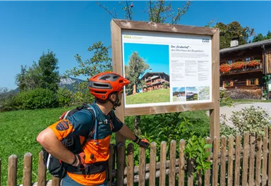 Ein Fahrradfahrer betrachtet ein Info-Schild in einer grünen Landschaft. Im Hintergrund sind traditionelle Holzhäuser und Bäume zu sehen.