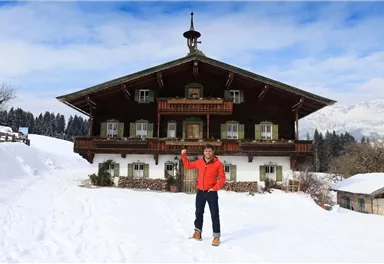Ein Holzhaus im alpinen Stil, umgeben von Schnee und Bergen. Eine Person steht davor und hebt die Hand.