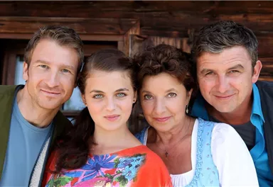 Eine Gruppe von vier Personen lächelt fröhlich in die Kamera. Sie stehen vor einem Holzhaus und tragen unterschiedliche, bunte Kleidung.