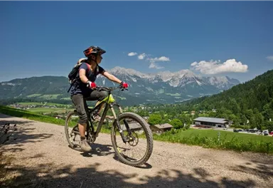 Eine Radfahrerin fährt auf einem schmalen Weg. Im Hintergrund sind Berge und eine grüne Landschaft zu sehen.