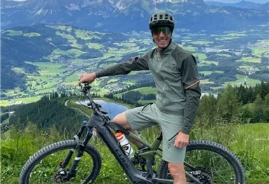 Ein Mountainbiker steht auf einem Weg mit Blick auf eine beeindruckende Berglandschaft. Die grüne Umgebung und der klare Himmel schaffen eine schöne Kulisse für das Abenteuer.