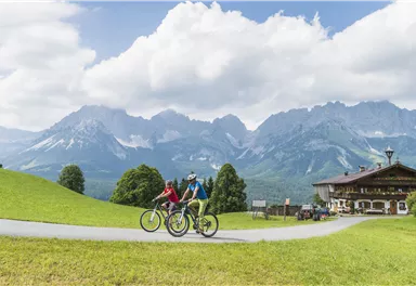 Zwei Radfahrer fahren auf einem Weg durch eine grüne Landschaft. Im Hintergrund sind majestätische Berge und ein traditionelles Gebäude zu sehen.