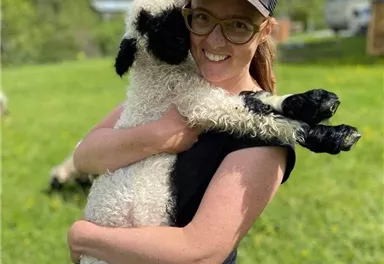 Eine Frau hält ein Lamm in den Armen und lächelt. Im Hintergrund sind Wiesen und Bäume zu sehen.