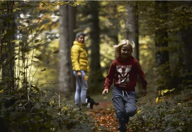 Ein Junge läuft fröhlich auf einem Waldweg, während eine Frau in warmer Kleidung ihn aus der Ferne beobachtet. Die Herbstfarben der Blätter schaffen eine gemütliche Atmosphäre.