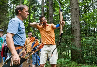 Eine Gruppe von Menschen übt das Bogenschießen im Wald. Ein Mann zielt mit einem Bogen, während ein anderer ihn beobachtet.