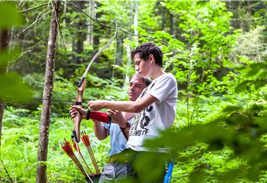 Zwei Personen stehen im Wald und üben mit einem Bogen. Der Trainer zeigt dem Schüler, wie man richtig zielt.