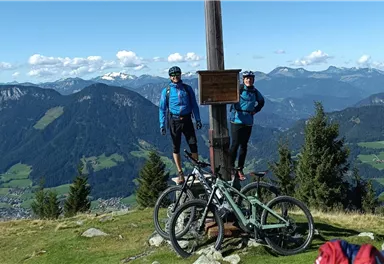 Zwei Radfahrer stehen an einem Gipfel mit einer Markierung. Im Hintergrund sind beeindruckende Berge und eine grüne Landschaft zu sehen.