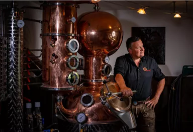 A man stands next to a copper distillation apparatus in a distillery. The surroundings are well-lit and display various machines and instruments.