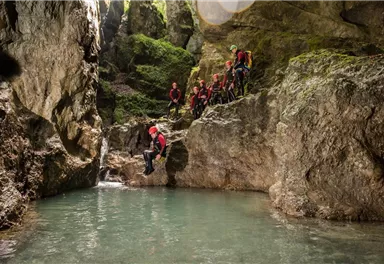Gruppen von Personen in roten Anzügen stehen an einem Felsen an einer kleinen Wasserstelle. Sie sind bereit zum Abseilen in den klaren, grünen Wasserpool.