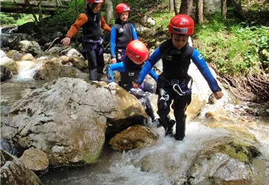 Vier Kinder mit Helmen und Neoprenanzügen waten durch einen kleinen Bach. Um sie herum sind Felsen und üppige grüne Bäume.