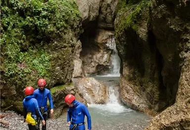 Eine Gruppe von Menschen in blauer Kleidung erkundet eine Schlucht mit Wasserfällen. Die Umgebung ist von grünen Felsen und Pflanzen umgeben.