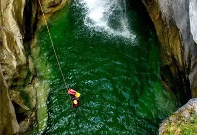 Ein Wasserfall fließt in ein tiefes grünes Becken. Darin schwimmen zwei Personen, während eine andere an einem Seil hinabsteigt.