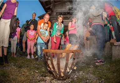 Eine Gruppe von Menschen um einen offenen Feuerkorb versammelt. Im Hintergrund ist ein Holzgebäude und leichter Rauch zu sehen.
