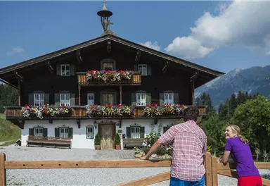 Ein traditionelles Holzhaus mit Balkonen, die mit bunten Blumen dekoriert sind. Im Hintergrund sind Berge und ein klarer Himmel zu sehen.